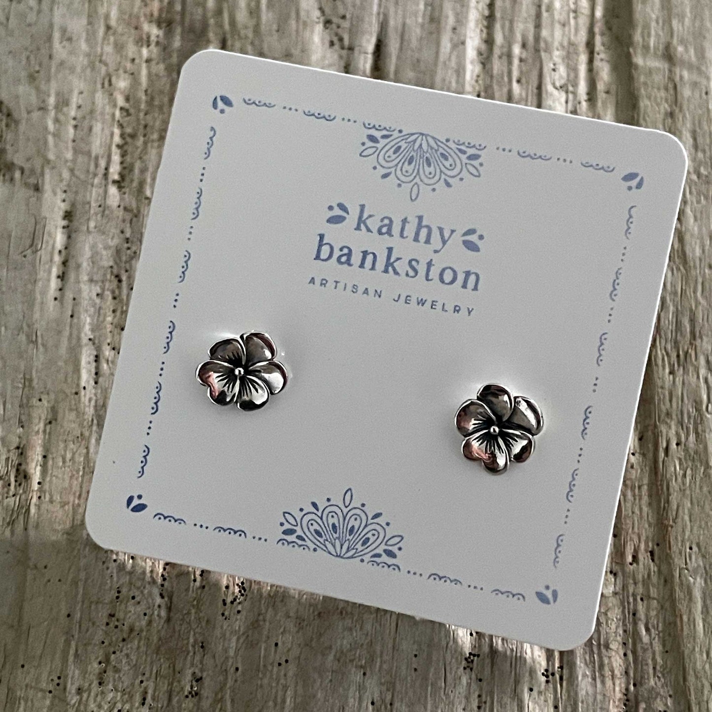 Flower-shaped earrings on a Kathy Bankston branded card against a wooden background