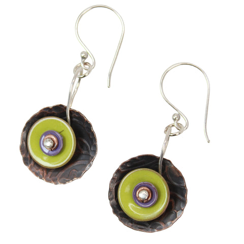 A pair of handmade dangle earrings featuring textured dark brown copper circular discs, a bright green enamel circle, a purple enamel ring, and a small silver bead at the center, suspended from sterling silver fishhook wires 