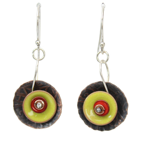 A pair of handmade dangle earrings featuring textured dark brown copper discs, bright lime green concentric circles, and a small red center, suspended from silver fishhook wires