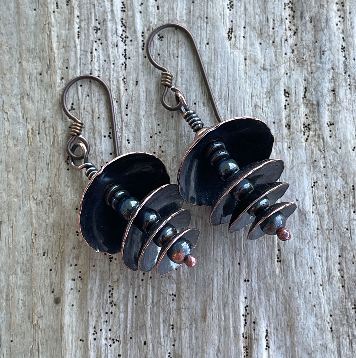 A pair of handmade drop earrings featuring dark copper discs and small beads, displayed on a weathered gray wood background.